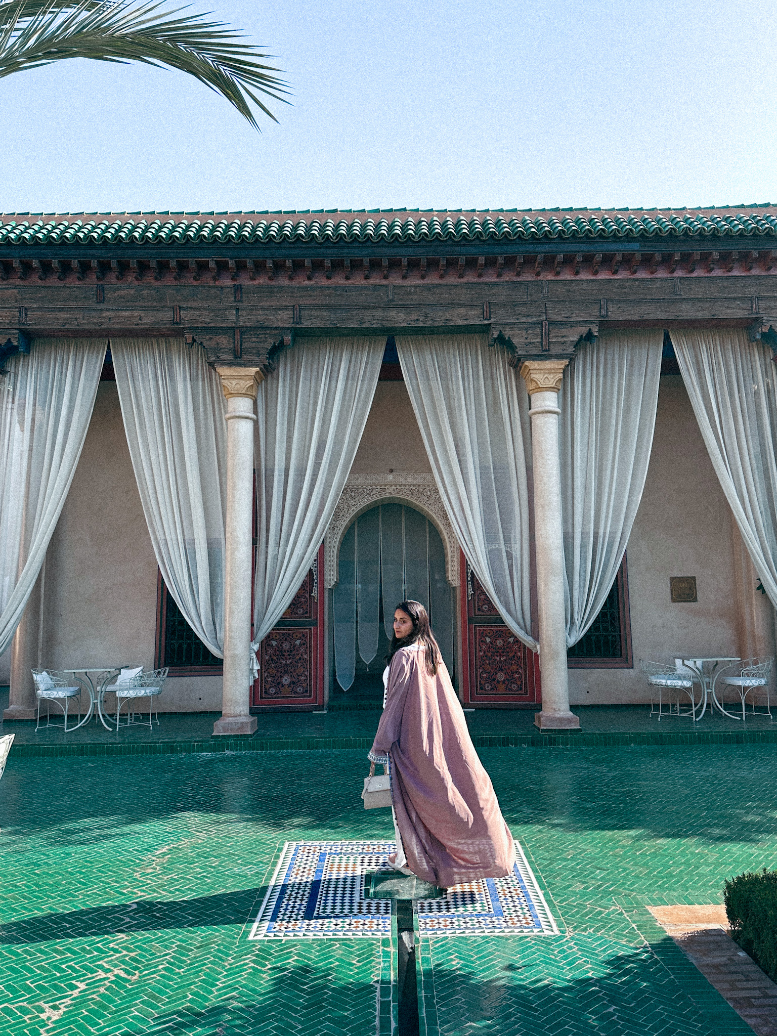 Inside le jardin secret marrakech. Green tiled floor. woman wearing pink abaya looking back. In front of her are draped curtains and a doorway.